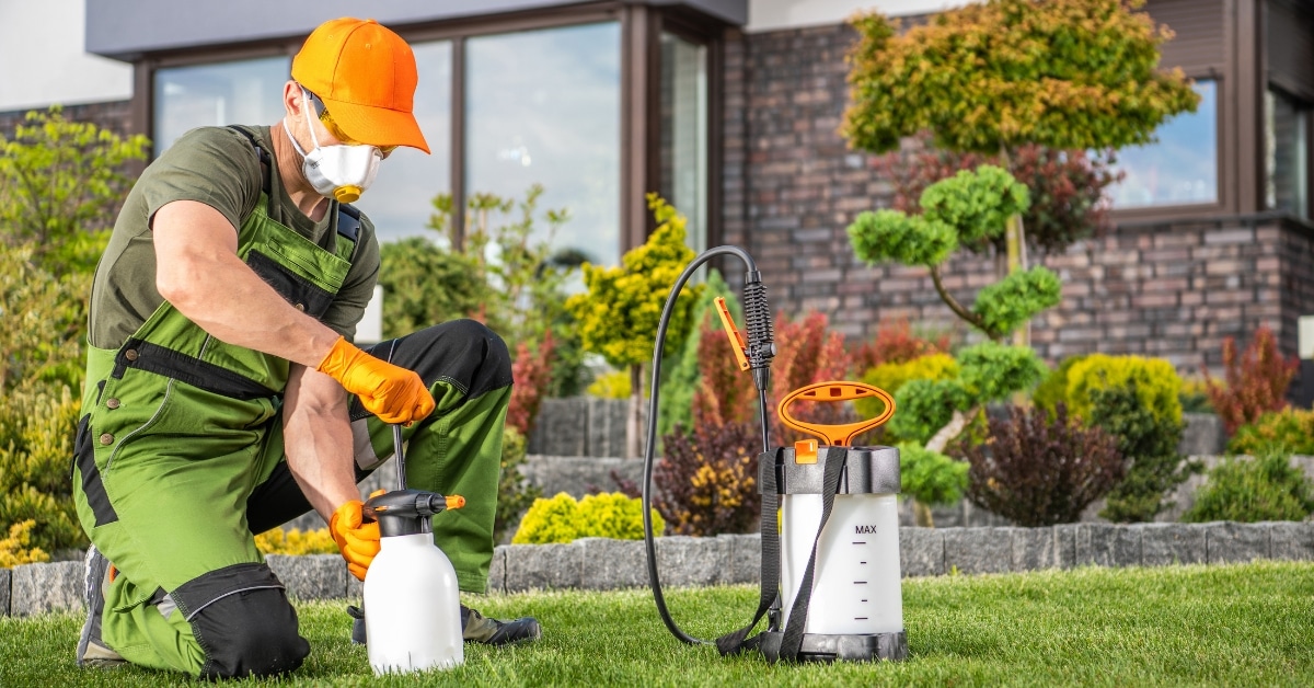 A worker in protective gear kneels on grass while holding a garden sprayer for pest control, with lush landscaping and plants visible in the background in Ogden, Utah.