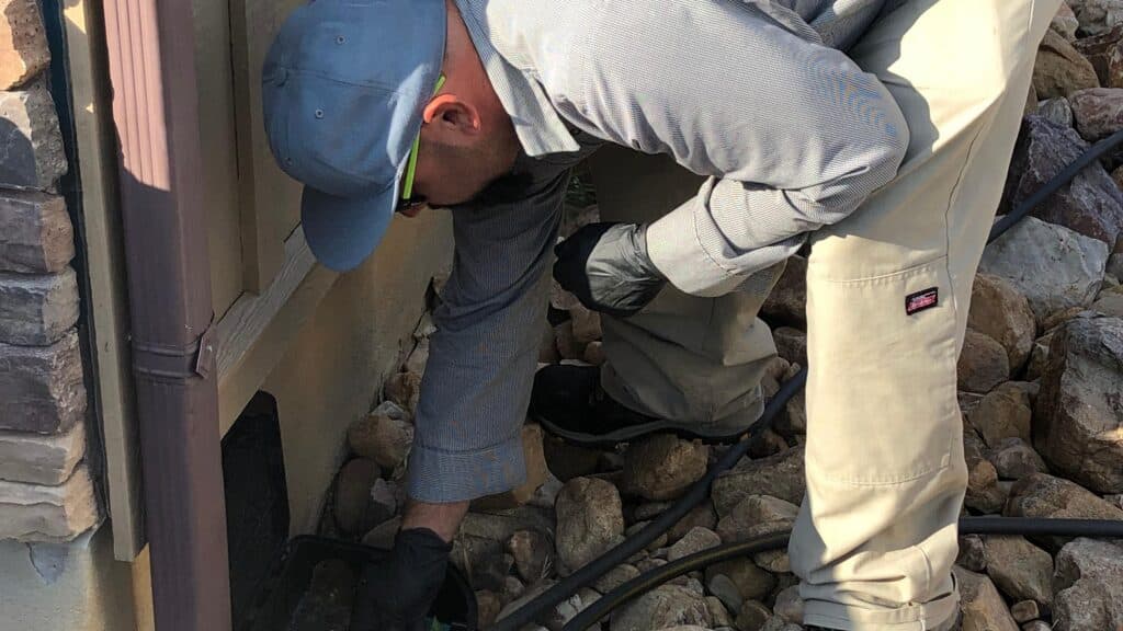 A person wearing gloves and work clothes bends down to inspect or work on an exterior wall near rocks and tubing, possibly performing Pest Control or Extermination Services in Plain City.