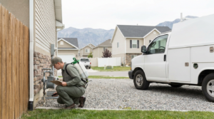 A pest control technician from West Point Utah inspects the exterior of a house, wearing a uniform and carrying equipment, with a white service van parked nearby in a suburban neighborhood.